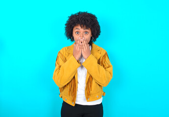 Young woman with afro hairstyle wearing yellow fringe jacket over blue background keeps hands on...