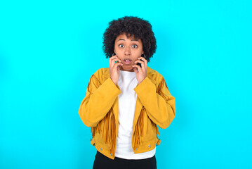 Speechless Young woman with afro hairstyle wearing yellow fringe jacket over blue background keeps...