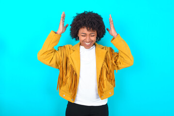 Young woman with afro hairstyle wearing yellow fringe jacket over blue background goes crazy as...