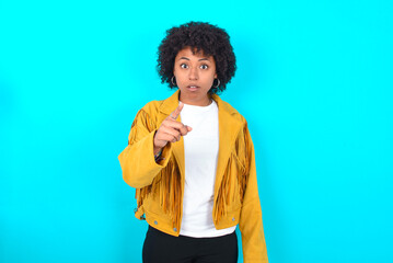 Shocked Young woman with afro hairstyle wearing yellow fringe jacket over blue background points at...