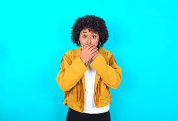 Stunned Young woman with afro hairstyle wearing yellow fringe jacket over blue background covers...