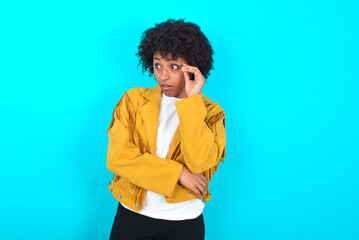 Astonished Young woman with afro hairstyle wearing yellow fringe jacket over blue background looks...