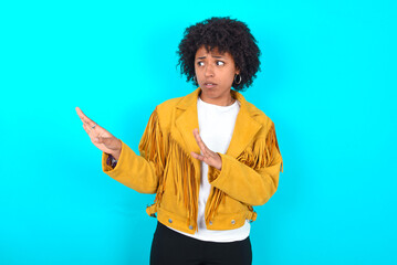 Displeased Young woman with afro hairstyle wearing yellow fringe jacket over blue background keeps...