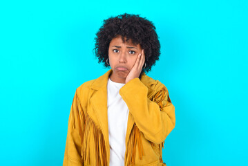 Fototapeta na wymiar Sad lonely Young woman with afro hairstyle wearing yellow fringe jacket over blue background touches cheek with hand bites lower lip and gazes with displeasure. Bad emotions