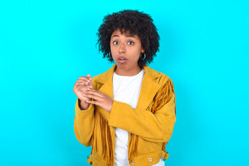 Surprised emotional Young woman with afro hairstyle wearing yellow fringe jacket over blue...