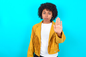 Young woman with afro hairstyle wearing yellow fringe jacket over blue background shows stop sign prohibition symbol keeps palm forward to camera with strict expression