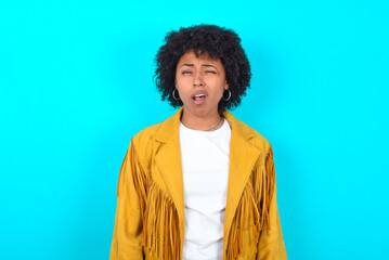 Fototapeta na wymiar Young woman with afro hairstyle wearing yellow fringe jacket over blue background yawns with opened mouth stands. Daily morning routine