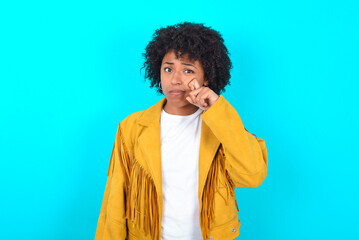 Fototapeta na wymiar Disappointed dejected Young woman with afro hairstyle wearing yellow fringe jacket over blue background wipes tears stands stressed with gloomy expression. Negative emotion