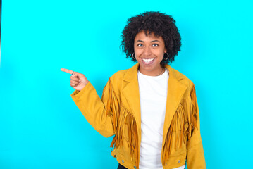 Young woman with afro hairstyle wearing yellow fringe jacket over blue background points aside on...