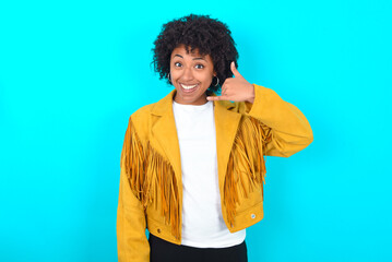 Young woman with afro hairstyle wearing yellow fringe jacket over blue background makes phone...