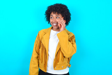 Fototapeta na wymiar Pleasant looking happy Young woman with afro hairstyle wearing yellow fringe jacket has nice telephone conversation and looks aside, has nice mood and smiles positively while talks via cell phone
