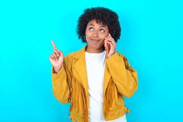 Young woman with afro hairstyle wearing yellow fringe jacket over blue background speaks on mobile...