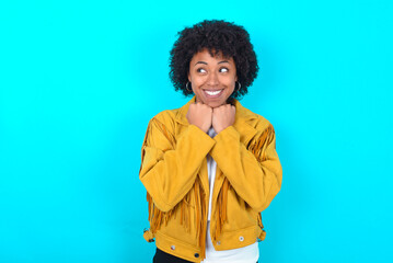 Obraz na płótnie Canvas Young woman with afro hairstyle wearing yellow fringe jacket over blue background holds hands under chin, glad to hear heartwarming words from stranger