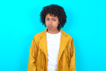 Young woman with afro hairstyle wearing yellow fringe jacket over blue background depressed and...