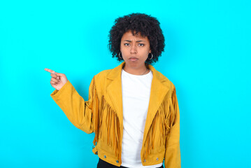Fototapeta na wymiar Serious Young woman with afro hairstyle wearing yellow fringe jacket over blue background smirks face points away on copy space shows something unpleasant. Look at this advertisement. Big price 