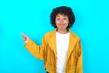 Obraz na płótnie Canvas Positive Young woman with afro hairstyle wearing yellow fringe jacket over blue background with satisfied expression indicates at upper right corner shows good offer suggests to click on link