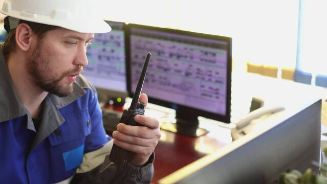 Serious Caucasian unshaven worker in overalls and helmet uses a walkie - talkie sitting in the control room . Automated workplace of a dispatcher or operator of a modern enterprise. Close up