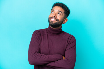 Young Brazilian man isolated on blue background looking up while smiling