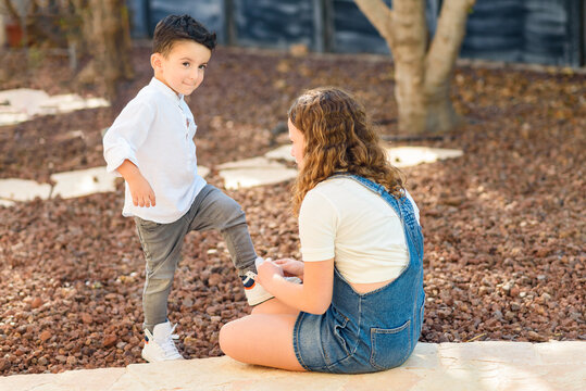 Teenager girl helps her little brother to tie shoelaces. Young babysitter and preschool kid spending time together, outdoor on summer day. Child in white shirt, denim, and sports shoes.