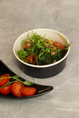 Vegetarian salad with potato, tomatoes, arugula and 
pumpkin seeds in round paper take-away container on gray background. Healthy food delivery concept. Free space for text.