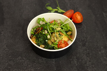 Vegetarian salad with potato, tomatoes, arugula and 
pumpkin seeds in round paper take-away container on dark background. Healthy food delivery concept. Free space for text.