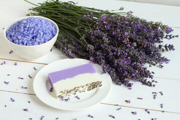 Obraz na płótnie Canvas Sea salt with lavender in a white bowl, soap made from lavender, olive oil and cocoa butter on a white plate and dried lavender flowers are located on a white wooden table. Closeup.