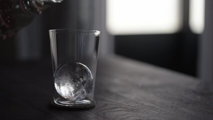 Slow motion pour tonic water over ice ball in tumbler glass on wood table - Powered by Adobe