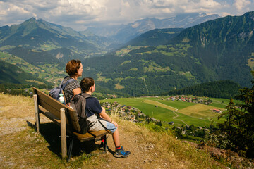 Fototapeta na wymiar Mother and son sitting on a bench enjoying amazing view high in the Vaud Alps while hiking on a sunny day in summer. Leysin, Switzerland
