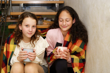 Two teenager girls sitting in the wood stairs wrapped in blanket with a cup of hot drink. Cold in home, problems with household heat supply in winter