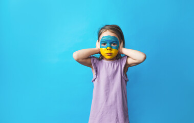 a little scared girl with a Ukrainian flag painted on her face covers her ears with her hands and so as not to hear anything on a blue background.