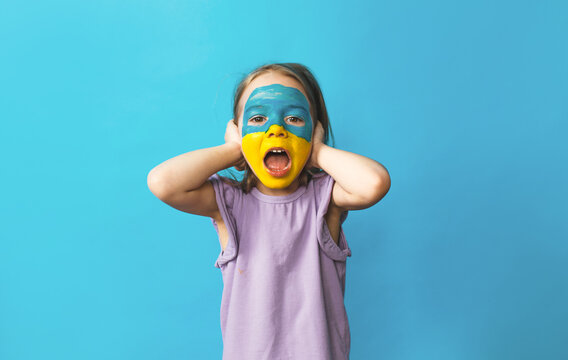a little scared girl with a Ukrainian flag painted on her face covers her ears with her hands and screams loudly so as not to hear anything - Powered by Adobe