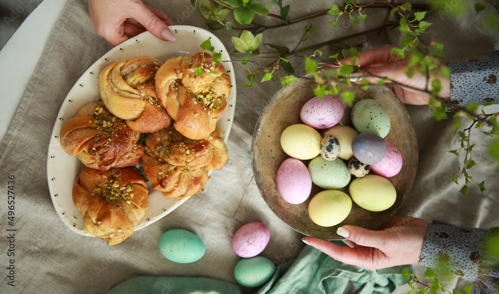 Wall mural Christian holiday, Easter. Easter buns with pistachios on a white plate, colored eggs with hands on the table. Tree branches, vases. Rustic. Background image, copy space. Top view