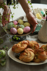 Christian holiday, Easter. Easter buns with pistachios on a white plate, colored eggs with hands on the table. Tree branches, vases. Rustic. Background image, copy space. Top view