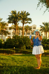 A young woman in a hat and a white skirt among the palm trees speaks on the phone while solving work issues.