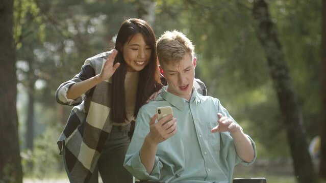 Being in the public park in a good weather, a young asian lady and her disabled blonde man finish talking with their friends
