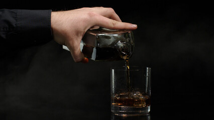 Hand of barman pouring golden whiskey, cognac or brandy from bottle into glass with ice cubes on black background. Bartender pouring glass of alcohol drink bourbon, rum. Copy-space