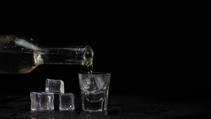 Shot glass on black table. Barman hand pours vodka, tequila or sake from bottle into empty shot glass on black background with ice cubes. Preparation to drink alcohol. Copy-space