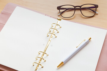 An open pastel pink leather planner with a pen and glasses on a wooden desk.