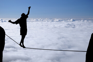 Silhouette of young man balancing on slackline high above clouds and mountains. Slackliner balancing on tightrope beautiful colorful sky and clouds behind, highline silhouette.