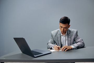 Asian man writing in a notebook laptop work at the desktop Gray background
