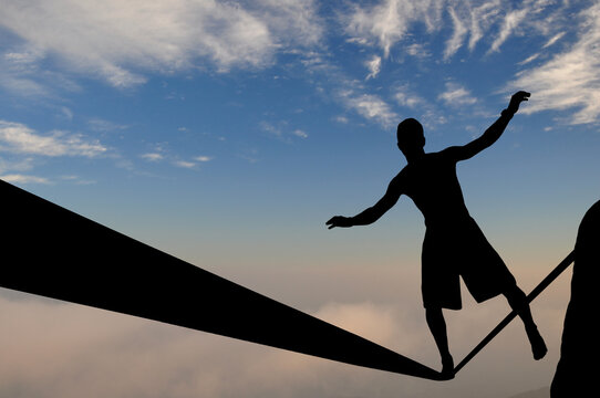 Silhouette of young man balancing on slackline high above clouds and mountains. Slackliner balancing on tightrope beautiful colorful sky and clouds behind, highline silhouette.