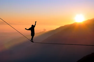 Zelfklevend Fotobehang Silhouette of young man balancing on slackline high above clouds and mountains. Slackliner balancing on tightrope during sunset, highline silhouette. © vp3k