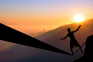 Silhouette of young man balancing on slackline high above clouds and mountains. Slackliner balancing on tightrope during sunset, highline silhouette.