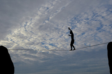 Silhouette of young man balancing on slackline, sun and clouds behind. Slackliner balancing on tightrope between two rocks, highline silhouette.