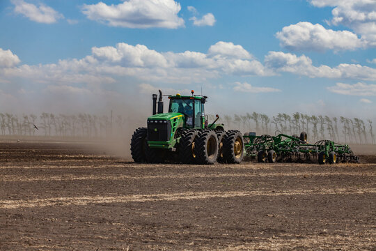 North Kazakhstan Province, Kazakhstan - May 12, 2012: Sowing campaign. John Deere tractor cultivating soil with plow. Dust cloud. Trees on background. Blue sky, clouds.