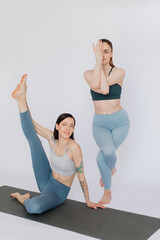 two female friends practice yoga asana poses on a mat on a white background. Beautiful slim girls are dressed in sportswear: blue leggings and tops