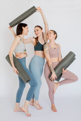 three beautiful girls smiling, laughing hold yoga mats in their hands in a white studio.
models are dressed in sports leggings and top. long shote