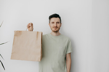 Closeup of Handsome young man over grey wall holding blank craft paper bag, mock-up of brown package and with white background