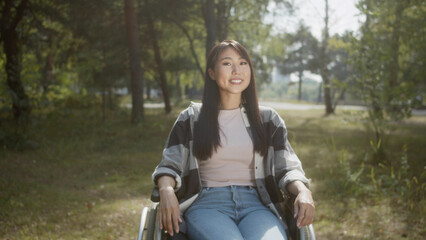 Fototapeta na wymiar An asian disabled female with long hair is posing, sitting in a wheelchair in a public park, smiling and touching hair