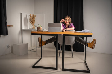 Young, tired, tortured, emotional, beautiful girl with glasses, works at work at the table, raised her legs relaxes, works remotely at home on the computer, remote work